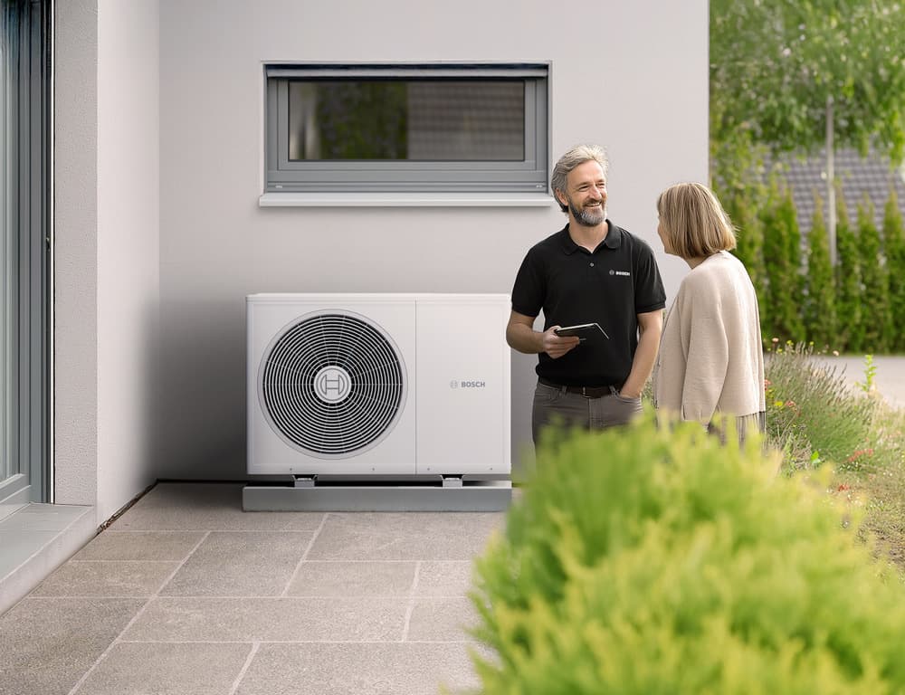 Technicien Bosch présentant une pompe à chaleur extérieure blanche à une cliente devant une maison.